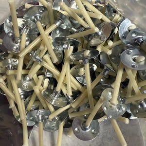 May include: A close-up shot of a pile of candle wicks with metal bases. The wicks are a light beige color, and the metal bases are silver. The wicks are arranged in a random pattern, with some wicks overlapping others. The wicks are for candle making.