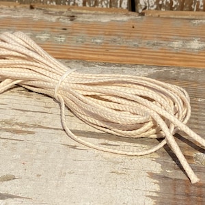 May include: A close-up of a spool of natural white cotton cord. The cord is braided and has a slightly textured surface. The cord is lying on a weathered wooden surface.