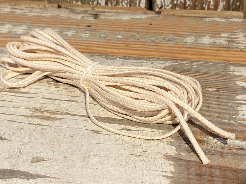May include: A close-up of a spool of natural white cotton cord. The cord is braided and has a slightly textured surface. The cord is lying on a weathered wooden surface.