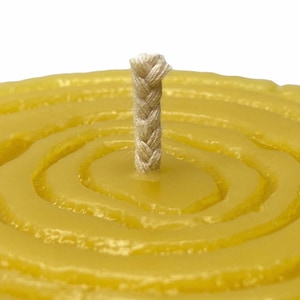 May include: A close-up of a round, yellow beeswax candle with a braided cotton wick in the centre. The candle has a textured, circular pattern.