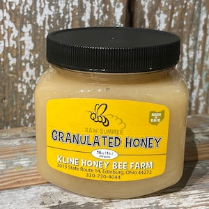 May include: A jar of granulated honey with a black lid and a yellow label. The label features a bee illustration and text that reads "RAW SUMMER GRANULATED HONEY" and "16 oz (1 lb) 454 grams". The jar is labeled "Made in Ohio".