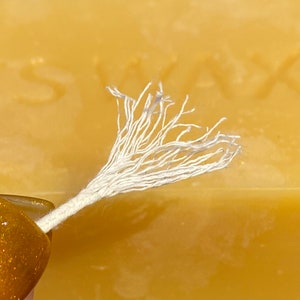 May include: Close-up of a white cotton candle wick with frayed ends. The wick is held over a block of yellow beeswax.
