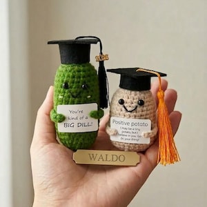 May include: Two crocheted graduation-themed figures: a green pickle and a tan potato, each wearing a black mortarboard cap with a tassel. The pickle holds a sign that reads "You're kind of a BIG DILL!" and the potato's sign says "Positive potato". A gold nameplate reads "WALDO".