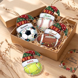 May include: Four flat acrylic ornaments shaped like sports balls including a soccer ball, baseball, football, and tennis ball. Each ball wears a green and red Christmas hat with gingerbread men and candy canes, personalized with names.