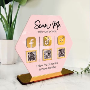 May include: Pink hexagonal sign with gold accents, featuring the text "Scan Me with your phone".  The sign includes icons for Facebook, TikTok, and Instagram, along with QR codes for each platform. The text "Follow me on socials & leave a review" is also included.