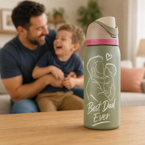 Op de afbeelding: Een groene waterfles met een witte en roze deksel. De fles heeft een witte afbeelding van een vader en kind met een hart, en de tekst "Best Dad Ever". De fles staat op een houten oppervlak.