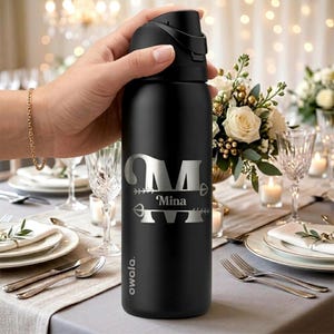 Puede incluir: Una botella de agua Owala de color negro mate con un gran monograma plateado de la letra M con un dise&ntilde;o de flecha y el nombre Mina impreso en el centro. La botella tiene una tapa negra con asa y se sostiene en posici&oacute;n vertical.