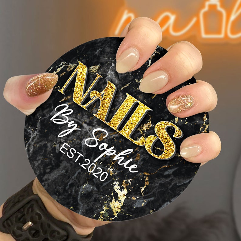 May include: A black and gold marble design round sticker with the text "NAILS by Sophie EST. 2020". A hand is holding the sticker, showing off a set of nude and glitter nail polish.