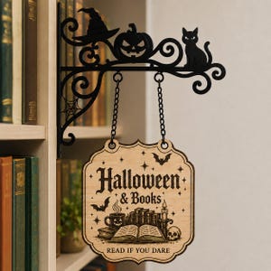 May include: A black metal wall bracket featuring a witch hat, pumpkin, and cat silhouette, suspending a light-colored wooden sign. The sign displays the text "Halloween & Books" and "READ IF YOU DARE" with illustrations of an open book, a mug, and a skull.