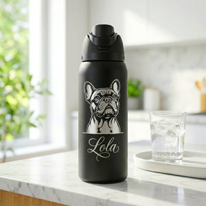 Puede incluir: Botella de agua aislada negra con tapa negra. Presenta una ilustraci&oacute;n de un bulldog franc&eacute;s y el nombre "Lola" en escritura cursiva. Un vaso de agua con hielo est&aacute; al lado.