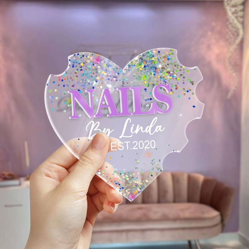May include: A clear heart-shaped acrylic sign with a glittery background. The sign says "NAILS by Linda EST. 2020" in purple lettering.