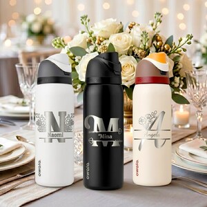 Puede incluir: Tres botellas de agua Owala con tapas abatibles. La botella blanca presenta una letra N floral y el nombre Naomi. La botella negra muestra una letra M con detalles de flechas y el nombre Mina. La botella crema muestra una letra A floral y el nombre Angela.