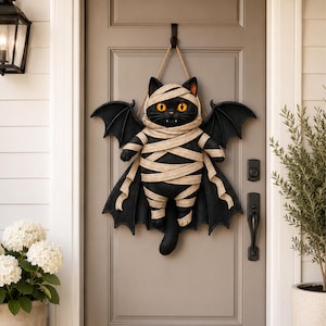 May include: A decorative wall hanging featuring a black cat wrapped in beige mummy bandages with large black bat wings. The cat has bright orange eyes and two small white fangs visible. It is suspended by a tan rope from a black metal hook.