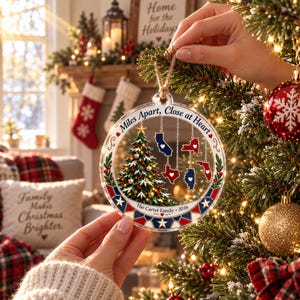 May include: A circular clear acrylic Christmas ornament featuring a printed Christmas tree, five state silhouettes with white hearts, and the text "Miles Apart, Close at Heart" and "The Carter Family 2026" with a red, white, and blue geometric border.