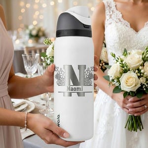 Puede incluir: Una botella de agua blanca Owala con tapa gris oscuro y asa. La botella presenta una letra N grande plateada con detalles florales y el nombre Naomi impreso en plata. El dise&ntilde;o est&aacute; centrado en la parte frontal de la botella cil&iacute;ndrica.