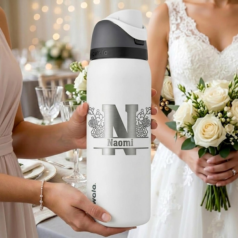 Puede incluir: Una botella de agua blanca Owala con tapa gris oscuro y asa. La botella presenta una letra N grande plateada con detalles florales y el nombre Naomi impreso en plata. El dise&ntilde;o est&aacute; centrado en la parte frontal de la botella cil&iacute;ndrica.
