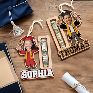 May include: Two wooden graduation money holders with personalized names "Sophia" and "Thomas". Each holder features a cartoon graduate with a graduation cap and gown, holding a diploma and a rolled-up dollar bill.