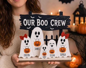 Puede incluir: Una decoración de Halloween de madera con cinco figuras de fantasmas blancos que sostienen cubos de calabaza naranjas. Los fantasmas tienen los nombres Evelyn, James, Sadie, Elijah y Sophia. Un letrero negro arriba dice Our Boo Crew.