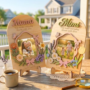 May include: Two wooden photo frames with floral and butterfly accents. One frame says "Mama" and the other says "Mimi". Each frame has a photo inside and the text "You make everyday bloom with love". The frames are on a wooden table.