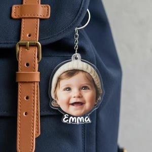 May include: A clear acrylic keychain featuring a smiling baby wearing a knit cap. The baby's image is in a circular shape, with the name "Emma" printed in white script below. The keychain is attached to a silver chain and a backpack.