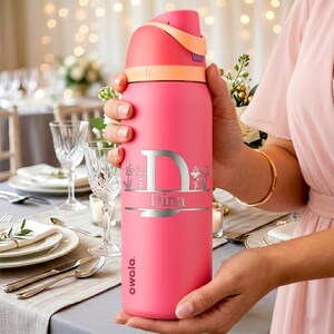 Puede incluir: Una botella de agua Owala de color rosa brillante con un dise&ntilde;o grabado en plata que muestra una letra D grande, el nombre Dina y peque&ntilde;as ilustraciones florales. La botella tiene una tapa color melocot&oacute;n con cierre abatible.
