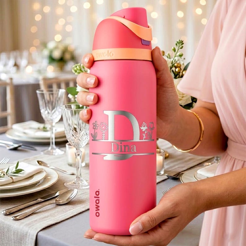Puede incluir: Una botella de agua Owala de color rosa brillante con un dise&ntilde;o grabado en plata que muestra una letra D grande, el nombre Dina y peque&ntilde;as ilustraciones florales. La botella tiene una tapa color melocot&oacute;n con cierre abatible.