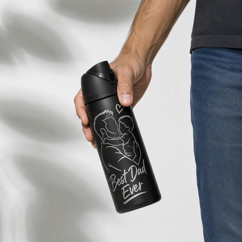 Op de afbeelding: Zwarte waterfles met een klapdeksel. De fles heeft een witte afbeelding van een vader en kind met de tekst "Best Dad Ever". De fles wordt door een persoon vastgehouden.