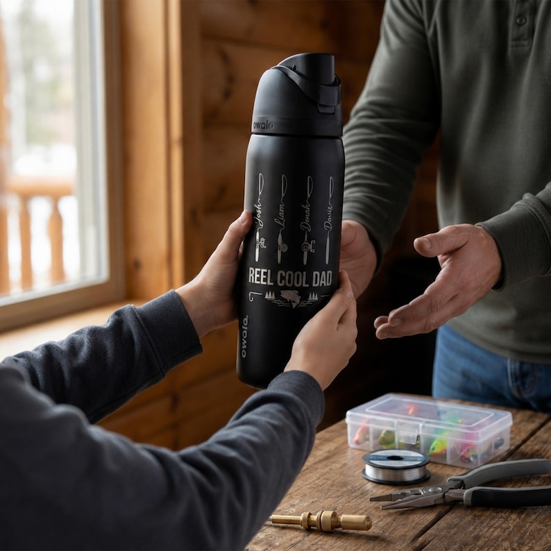 May include: A matte black Owala water bottle featuring a white graphic of four fishing rods with names and the text REEL COOL DAD printed on the front. The bottle has a black lid with a carry loop and a push-button opening mechanism.