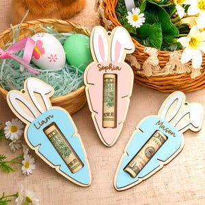 May include: Wooden Easter carrot-shaped gift holders in pink and blue, each with a name and a rolled-up bill inside. The carrots have bunny ear accents and are displayed near Easter eggs and baskets.