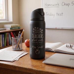 May include: A black Owala water bottle with a flip-top lid. The bottle features the text "TEACHERS plant seeds THAT GROW FOREVER" and "Mrs. Smith" in a floral design. An apple, books, and school supplies are on the desk.