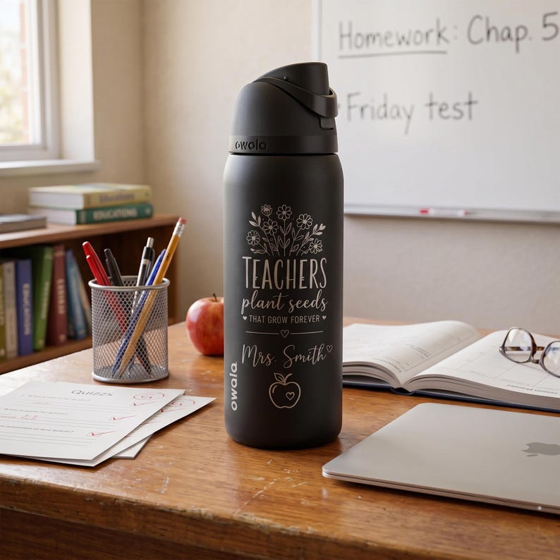 May include: A black Owala water bottle with a flip-top lid. The bottle features the text "TEACHERS plant seeds THAT GROW FOREVER" and "Mrs. Smith" in a floral design. An apple, books, and school supplies are on the desk.