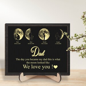 May include: A black framed print with a gold background featuring four moons with silhouettes of a father and child. The moons represent the dates of birth of the father and his children. The text reads "Dad, The day you became my dad this is what the moon looked like, We love you!"