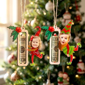 May include: Two wooden Christmas ornaments hanging by twine, each featuring a rectangular frame holding a rolled-up dollar bill. One ornament shows a child's photo with the name Emily, and the other shows a child's photo, both dressed as elves.