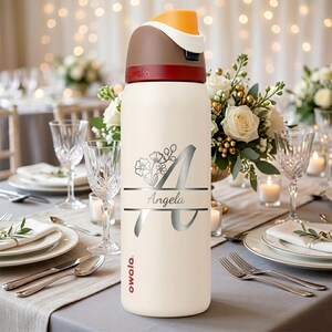 Puede incluir: Una botella de agua Owala de color crema con tapa marr&oacute;n oscuro y un detalle naranja. La botella est&aacute; personalizada con una letra A grande, un dise&ntilde;o floral y el nombre Angela impreso en una fuente cursiva oscura en la parte frontal.
