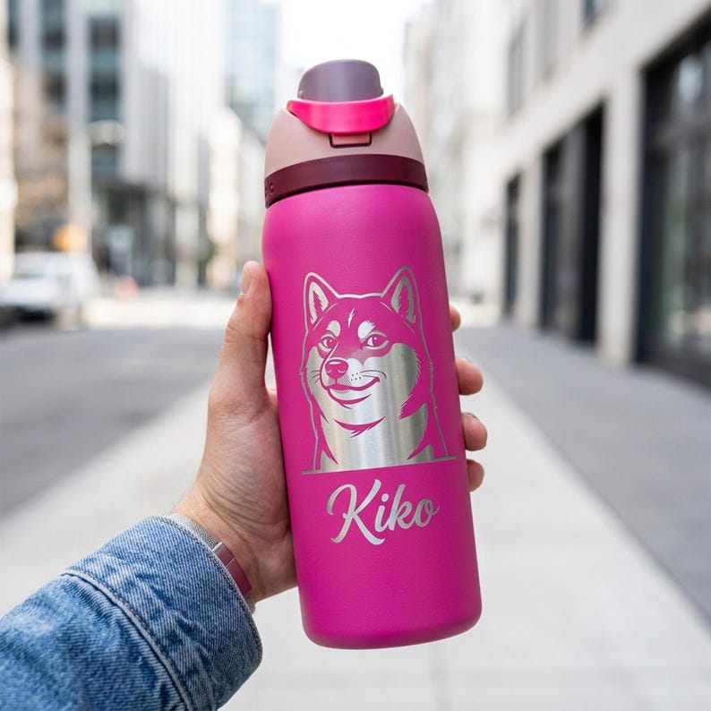 Puede incluir: Una botella de agua rosa con un dise&ntilde;o de perro Shiba Inu plateado y el nombre "Kiko" en cursiva. La botella tiene una tapa rosa oscuro y una tapa rosa claro. La botella est&aacute; sujeta en la mano de una persona.
