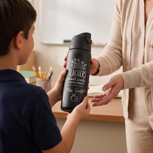 May include: A black insulated water bottle with a flip-top lid. The bottle features the text "TEACHERS plant seeds that grow forever" and "Mrs. Smith" with an apple graphic. The bottle is being handed to a person.