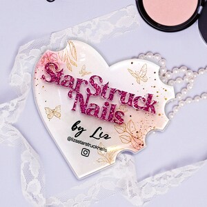 Op de afbeelding: Een hartvormig acryl bord met de tekst "Starstruck Nails by Liz" in roze glitterletters. Het bord heeft een aquarel achtergrond met gouden vlinders en bloemmotieven. Het Instagram logo is ook inbegrepen.