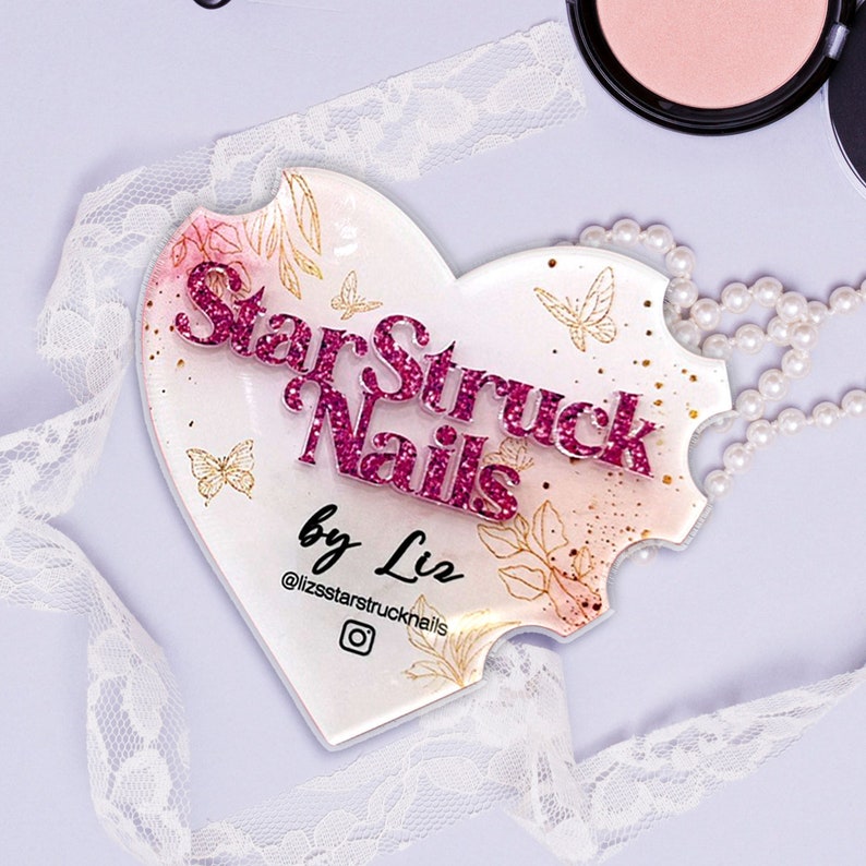 Op de afbeelding: Een hartvormig acryl bord met de tekst "Starstruck Nails by Liz" in roze glitterletters. Het bord heeft een aquarel achtergrond met gouden vlinders en bloemmotieven. Het Instagram logo is ook inbegrepen.