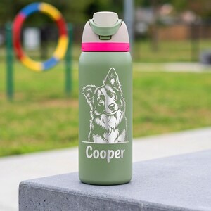 Puede incluir: Una botella de agua aislada verde con un retrato de perro blanco y el nombre "Cooper" impreso debajo. La botella tiene una tapa blanca y verde con un acento rosa. El fondo es una escena borrosa del parque.