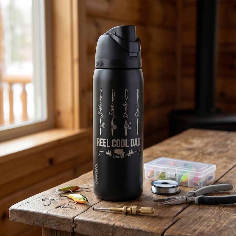 May include: A matte black Owala water bottle with a black lid, featuring white laser-engraved graphics of four fishing rods with the names Josh, Liam, Jonah, and Davis, and the text REEL COOL DAD above a fish icon and pine trees.