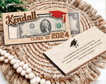 May include: Wooden graduation money holder with a slot for a dollar bill. The holder is decorated with a graduation cap, the name Kendall, and the text "Class of 2024". A separate wooden card with a graduation cap and a message of congratulations is also included.