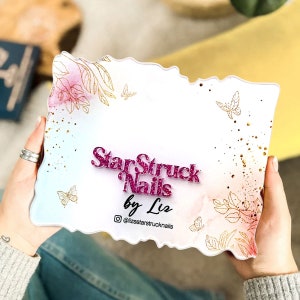 Op de afbeelding: Een wit bord met een roze en blauwe aquarel achtergrond en gouden glitter accenten. Het bord zegt "StarStruck Nails by Liz" met een roze glitter lettertype en een gouden glitter Instagram-handvat "@lizsstarstrucknails".