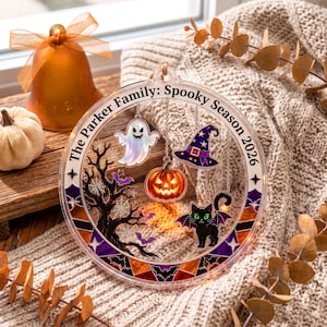 May include: A circular acrylic Halloween ornament featuring the text The Parker Family: Spooky Season 2026. It includes hanging charms of a ghost, a jack-o'-lantern, a witch hat, and a black cat with purple wings, set against a tree and web design.