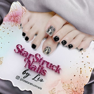 Op de afbeelding: Close-up van iemands voeten met zwarte en glitternagellak. De teennagels zijn versierd met een slangenhuidpatroon. De afbeelding bevat de tekst "StarStruck Nails by Liz @lizsstarstrucknails".