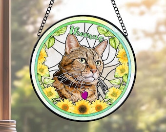 May include: Round stained glass cat suncatcher with a brown tabby cat portrait, surrounded by sunflowers and green leaves. The cat has a purple heart-shaped tag. The name "Thomas" is written in green script at the top. The suncatcher hangs from a black chain.