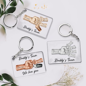 May include: Three clear acrylic keychains with silver key rings. Each keychain features a hand with intertwined fingers, representing a family. The hands are drawn in black and brown ink. The keychains have the text "Daddy's Team" and include the names of family members. One keychain also includes the text "We love you".