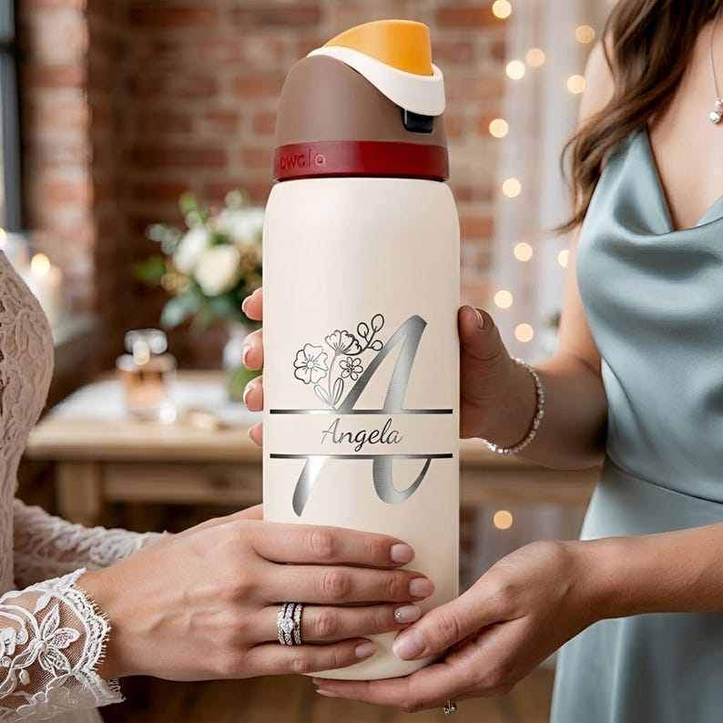 Puede incluir: Una botella de agua de color crema con una tapa de color marr&oacute;n oscuro y rojo con un acento naranja. La botella est&aacute; personalizada con una letra A grande de color plateado, un gr&aacute;fico floral y el nombre Angela en letra cursiva en el centro.