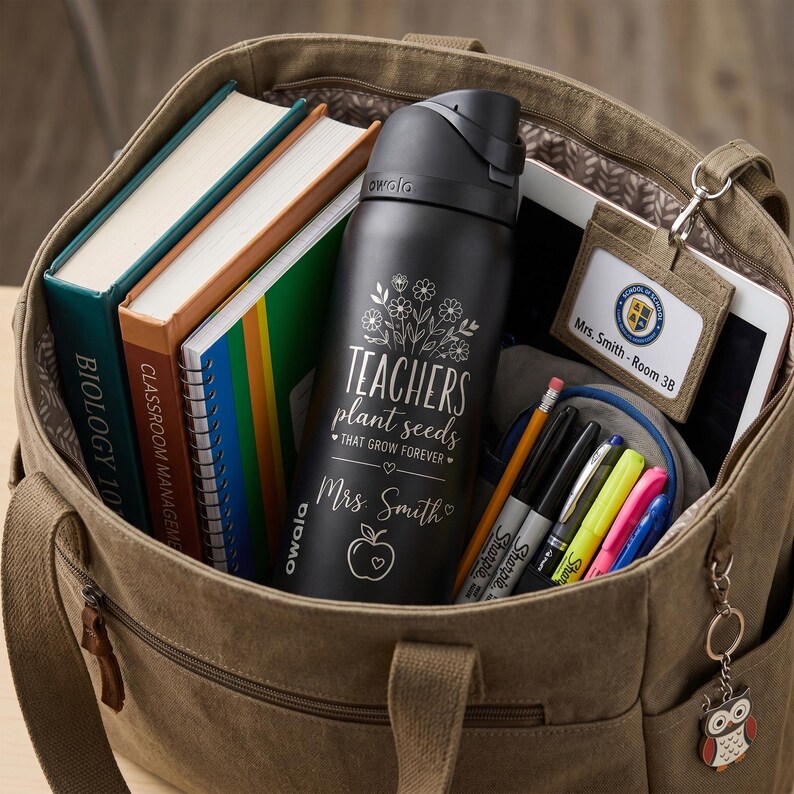 May include: A brown canvas tote bag filled with school supplies. Inside are books, a black water bottle with the text "TEACHERS plant seeds that grow forever", a tablet, and a pencil case with markers.