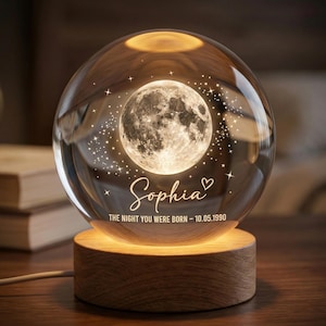 May include: A clear glass sphere with a detailed moon design and star accents, illuminated by a warm light. The sphere sits on a wooden base, with the name "Sophia" and a date inscribed on the glass.