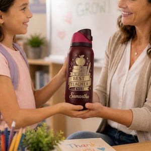 May include: A maroon water bottle with a gold design featuring an apple, books, and a pencil. The text reads "BEST TEACHER EVER" and "Samantha". The bottle has a dark red lid and is being held by two people.